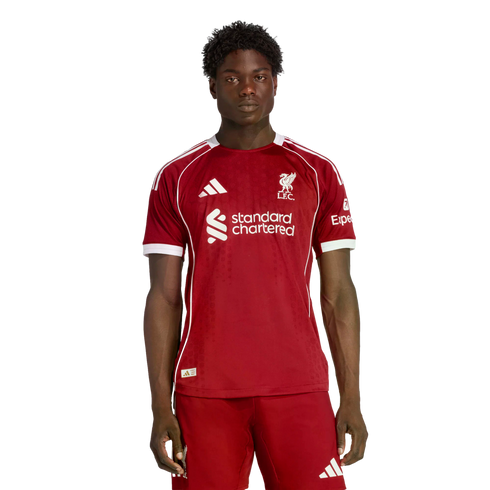 A young man wearing the red 2024-25 Liverpool F.C. home soccer kit, standing against a black background.
