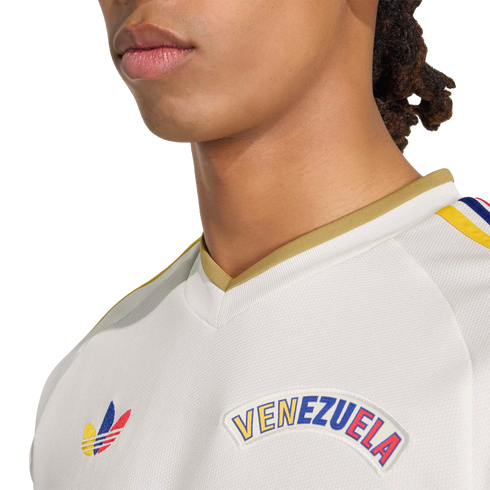 Close-up of a person wearing a white Adidas Venezuela jersey with a gold collar and colorful logos on the chest, set against a black background.