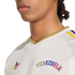 Close-up of a person wearing a white Adidas Venezuela jersey with a gold collar and colorful logos on the chest, set against a black background.