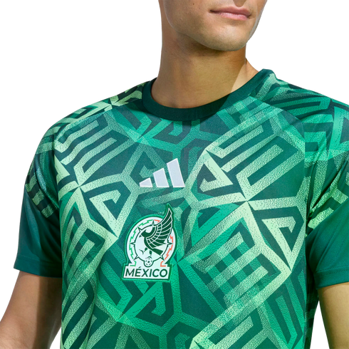 A man wears a green Adidas Mexico soccer jersey featuring a bold, geometric pattern inspired by ancient civilizations and the official team crest.