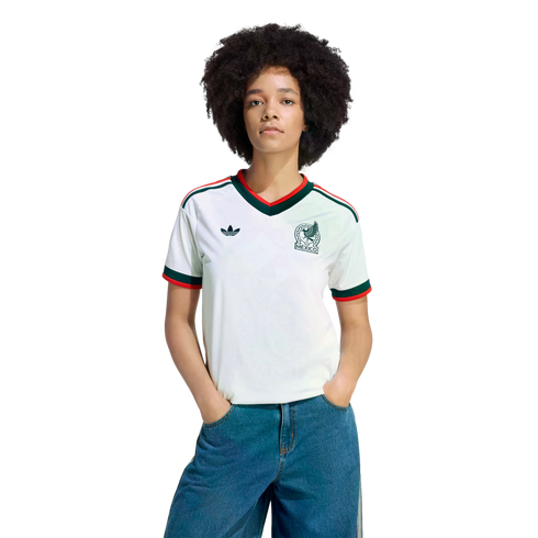 A person with dark curly hair wears a white Mexico Adidas soccer jersey with green and red trim and blue jeans against a solid black background.