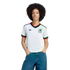 A person with dark curly hair wears a white Mexico Adidas soccer jersey with green and red trim and blue jeans against a solid black background.