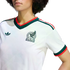 A close-up shot of a person from the chest up, wearing a white Mexico national team soccer jersey by Adidas. The jersey features a green and red V-neck collar, green and red stripes on the shoulders, and matching cuffs on the short sleeves. The Mexico nat
