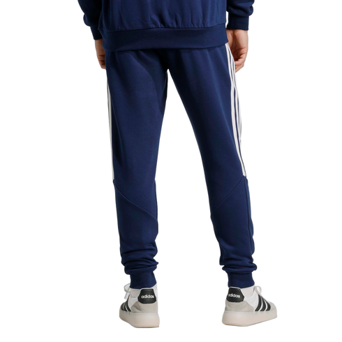 Rear view of a person wearing navy Adidas track pants with white side stripes and white sneakers against a light gray background.