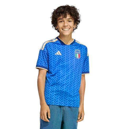 A smiling boy with curly hair wearing a blue patterned Italy soccer jersey and blue jeans, standing against a black background.