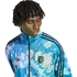A person with short dreadlocks wearing a blue and white tie-dye patterned Adidas track jacket with a San Jose Earthquakes logo.