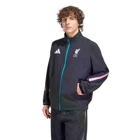 A person with curly brown hair models a black Adidas Liverpool F.C. track jacket featuring teal and pink accents, standing against a solid black background.