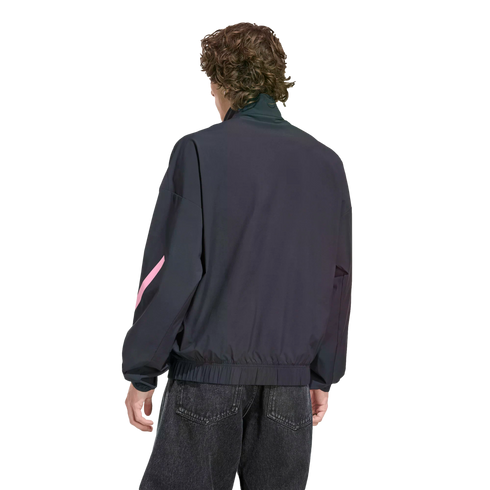 Rear view of a person with wavy brown hair wearing a dark navy zip-up jacket with a pink design on the sleeve and black jeans, set against a black background.