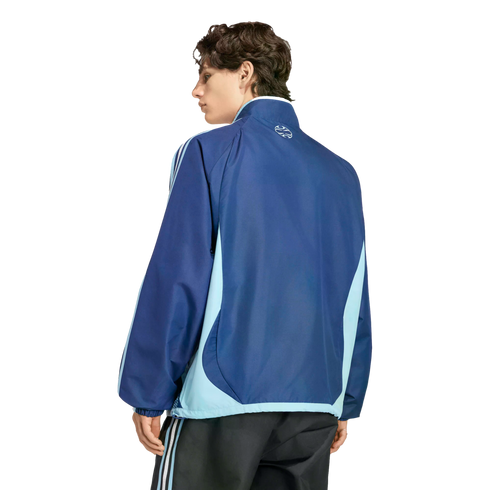 Back view of a man wearing a navy and light blue color-blocked track jacket and dark striped pants, looking over his shoulder against a black background.