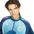 A young man with wavy dark hair and blue eyes wears a blue Argentina national football team track jacket, set against a solid black background.
