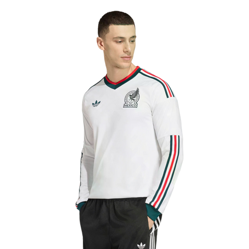 A man wearing a white long-sleeved Adidas Mexico soccer jersey with green and red trim, paired with black tracksuit pants, against a black background.