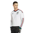 A man wearing a white long-sleeved Adidas Mexico soccer jersey with green and red trim, paired with black tracksuit pants, against a black background.
