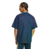 Back view of a man with braided hair wearing a dark blue t-shirt with a geometric pattern and navy shorts with yellow stripes against a solid black background.