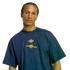 A man wears a dark blue t-shirt with yellow Adidas and Arsenal logos. The shirt features an all-over geometric pattern, and the background is solid black.