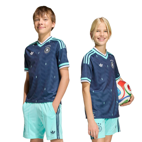 A boy and a girl smile while wearing matching navy blue and light blue Adidas soccer kits. The girl holds a soccer ball against a solid black background.