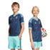 A boy and a girl smile while wearing matching navy blue and light blue Adidas soccer kits. The girl holds a soccer ball against a solid black background.