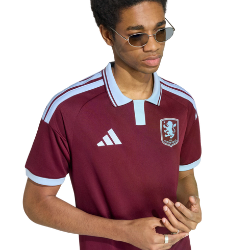 A person with sunglasses and rings wears a maroon and light blue Adidas Aston Villa polo shirt against a plain white background.