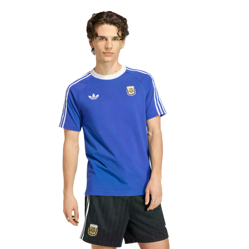 A young man with wavy brown hair models a royal blue Adidas Argentina national football team t-shirt and matching black athletic shorts against a solid black background.