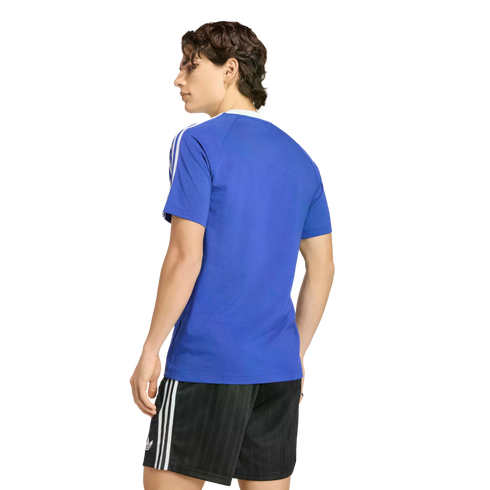 A rear view of a person with wavy brown hair wearing a blue t-shirt with white stripes on the sleeves and black athletic shorts with white stripes, standing against a solid black background.
