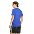 A rear view of a person with wavy brown hair wearing a blue t-shirt with white stripes on the sleeves and black athletic shorts with white stripes, standing against a solid black background.