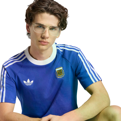 A young man with dark, wavy hair and gold-rimmed glasses wearing a blue Adidas Argentina national football team t-shirt against a black background.