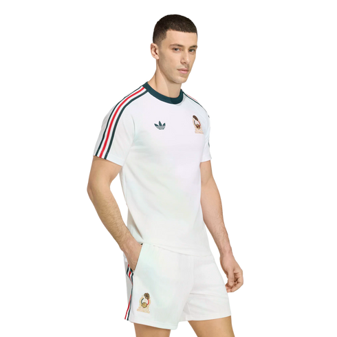 A man models a white Adidas football kit with green and red striped accents on the sleeves and shorts, featuring a gold team crest, against a solid black background.