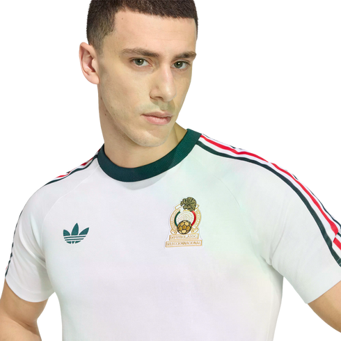A close-up of a young man wearing a white retro Mexico National Team t-shirt by Adidas. The shirt features a dark green crew neck, a green Adidas Trefoil logo on the right, and the Mexican National Team crest on the left. Three stripes in green, white, an