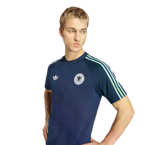 Young man with short blonde hair wearing a navy blue Adidas Germany t-shirt with mint green stripes, looking down and to the side against a black background.