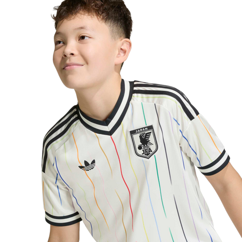 A young person smiling in a white Adidas Japan football jersey with vertical rainbow stripes, set against a solid black background.