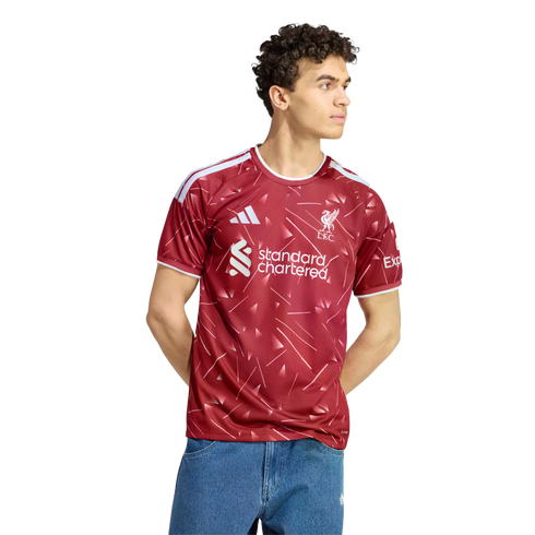 A man with curly hair wearing a red and white Liverpool FC soccer jersey and blue jeans, standing against a white background.