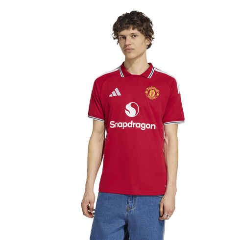 A man wearing a red Manchester United home football jersey with Snapdragon sponsorship and Adidas branding, paired with blue jeans, against a plain white background.