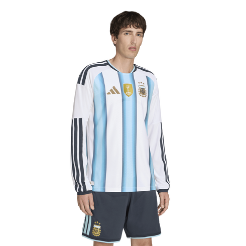 A man wearing an Argentina long-sleeve soccer jersey with light blue stripes and gold accents, and matching dark grey shorts.