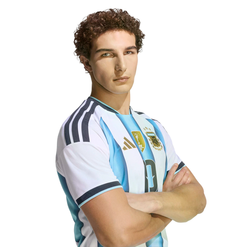 A man with curly brown hair wearing an Argentina national football team jersey, posing with arms crossed against a black background.