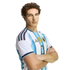 A man with curly brown hair wearing an Argentina national football team jersey, posing with arms crossed against a black background.