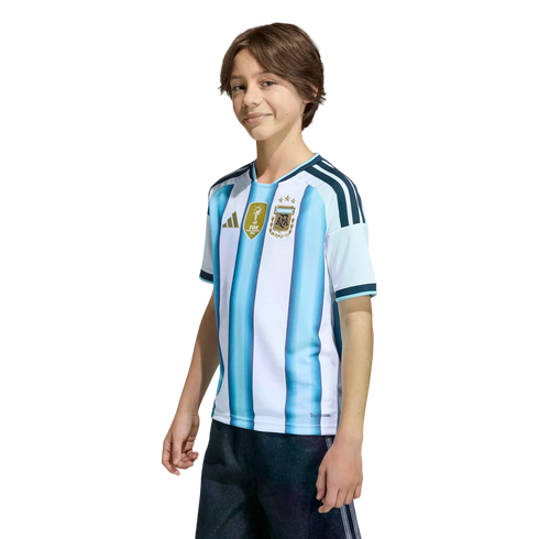 A smiling boy wearing an Argentina national football team jersey with the FIFA World Champions 2022 patch, set against a black background.