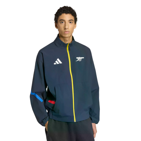 A man wears a navy blue Adidas track jacket with a yellow zipper, featuring a white Adidas logo and the Arsenal FC cannon crest.