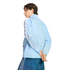 A man in a light blue zip-up track jacket and denim jeans looks back over his shoulder against a black background.