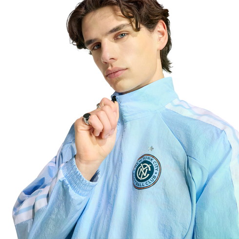 A man with dark wavy hair wears a light blue New York City Football Club zip-up jacket, posing against a black background.