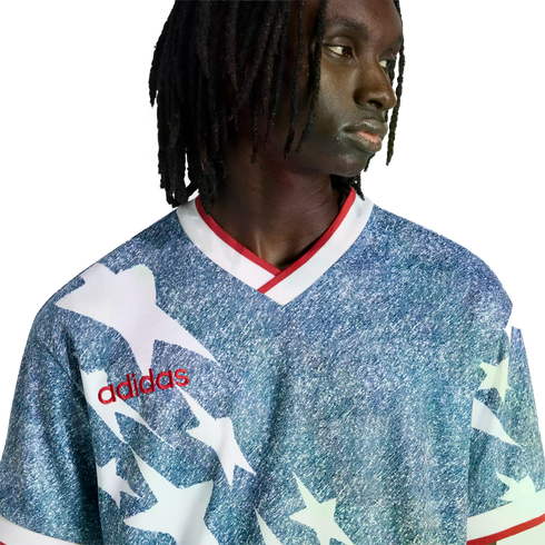 A person with dreadlocks wears a blue and white star-patterned Adidas jersey with a red logo and collar, set against a solid black background.