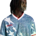 A person with dreadlocks wears a blue and white star-patterned Adidas jersey with a red logo and collar, set against a solid black background.