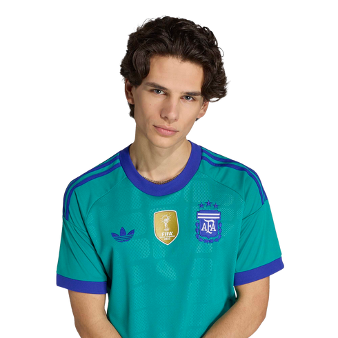 Portrait of a young man with wavy brown hair wearing a teal Argentina national team jersey with purple details and a gold FIFA patch against a black background.