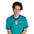 Portrait of a young man with wavy brown hair wearing a teal Argentina national team jersey with purple details and a gold FIFA patch against a black background.