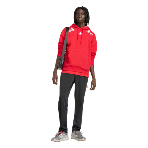 A Black person with dreadlocks stands against a black background in a red hoodie, black track pants, and grey sneakers, with a black crossbody bag.