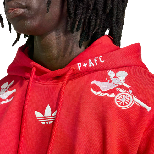 Close-up of a person in a red Adidas x Arsenal hoodie with white embroidered cherub riding a cannon, a Trefoil logo, and "P+AFC" text on the hood.