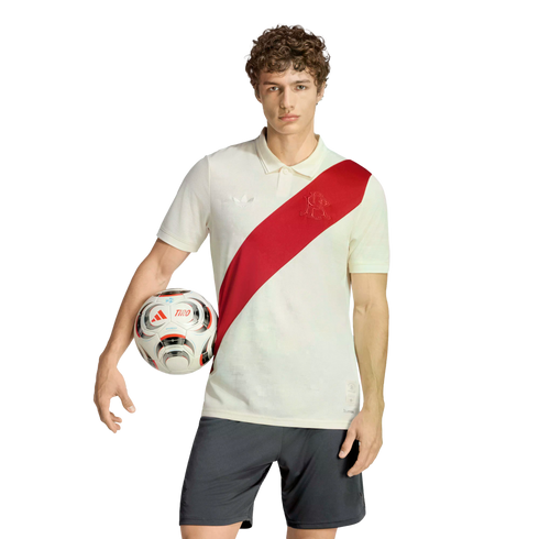 A man with curly hair stands against a white background, wearing an off-white polo with a red diagonal sash and dark shorts, holding a soccer ball under his arm.