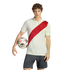 A man with curly hair stands against a white background, wearing an off-white polo with a red diagonal sash and dark shorts, holding a soccer ball under his arm.