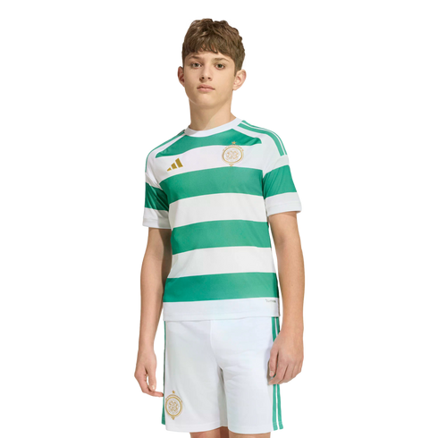 A young person modeling a Celtic FC soccer kit with green and white hoops, gold logos, and white shorts against a plain white background.