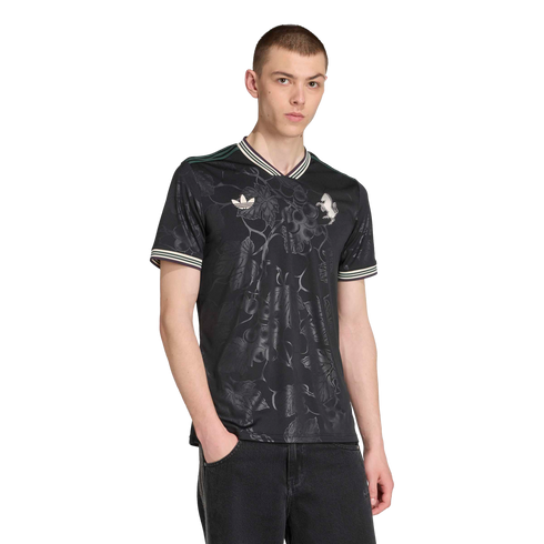A male model wearing a black floral-patterned Adidas jersey and dark jeans against a solid black background.