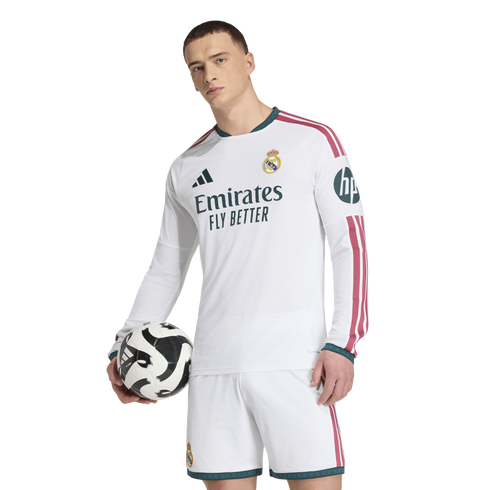 A man in a white Real Madrid long-sleeve jersey and shorts holds a soccer ball against a plain white background.