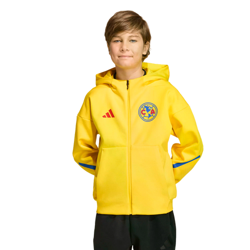 A boy wearing a yellow Club América adidas zip-up hoodie and black pants against a black background.
